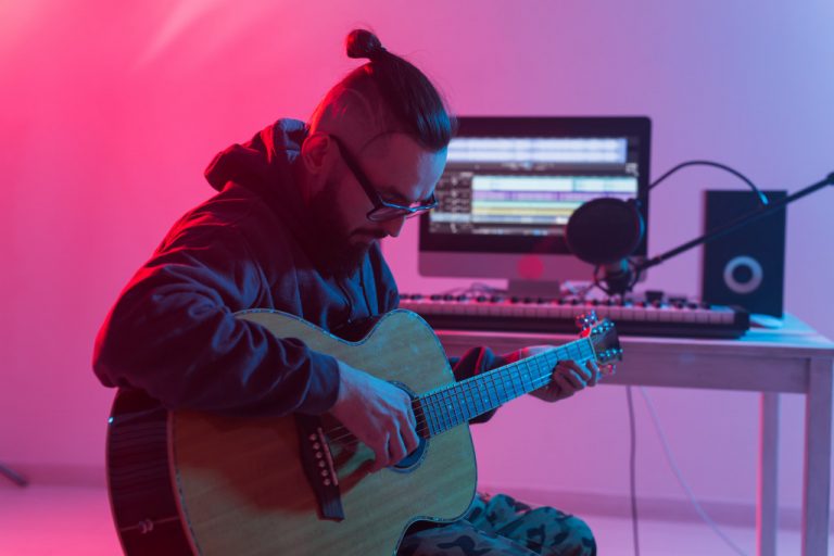 An indie artist plays the guitar at their home studio in front of a keyboard, mic, and computer.